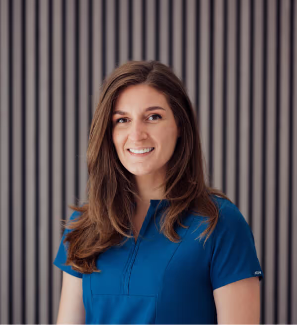 Smiling woman with long brown hair wearing a blue top standing against a gray vertical striped background.