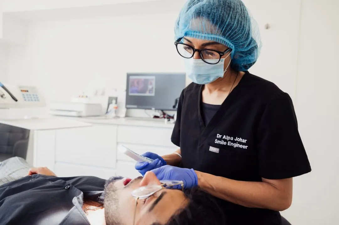 Dentist wearing surgical mask, hairnet, and gloves examining a male patient's mouth in a dental clinic.
