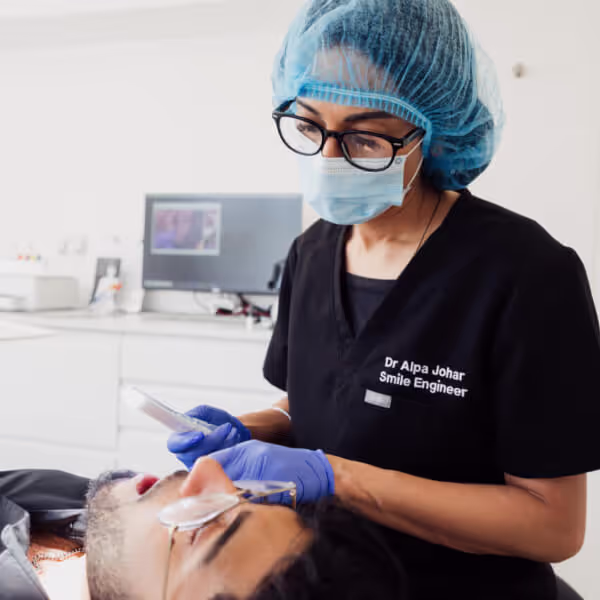 Dental professional wearing a blue hairnet, mask, glasses, and gloves working on a patient's teeth in a clinical setting.
