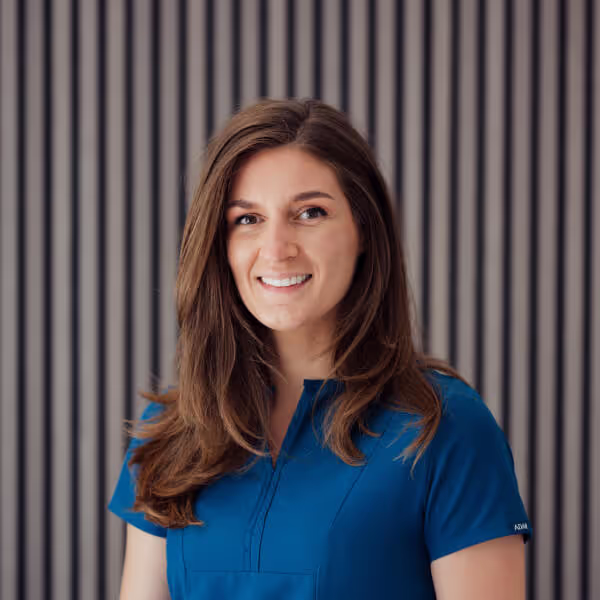 Smiling woman with long brown hair wearing a blue medical scrub top standing in front of a striped gray background.