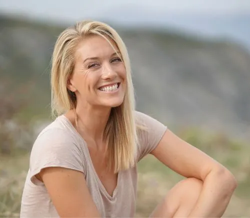 Smiling blonde woman with a light beige shirt sitting outdoors with a blurred natural background.