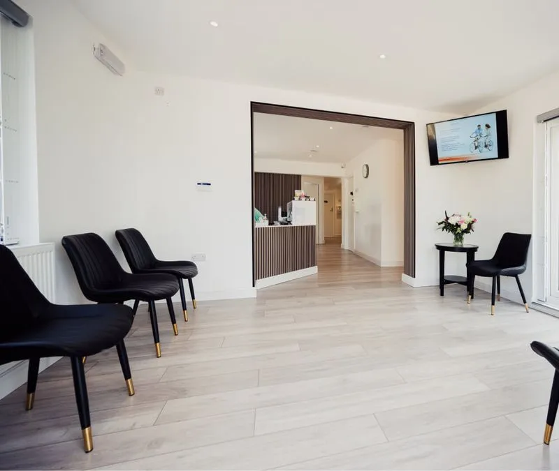 Bright waiting room with light wood flooring, black chairs with gold-tipped legs, a small table with a flower vase, and a wall-mounted TV.