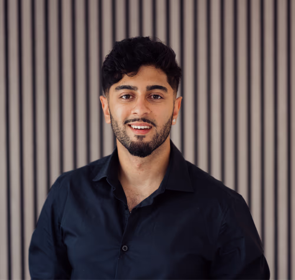 Smiling man with dark hair and beard wearing a black shirt standing against a vertically striped background.