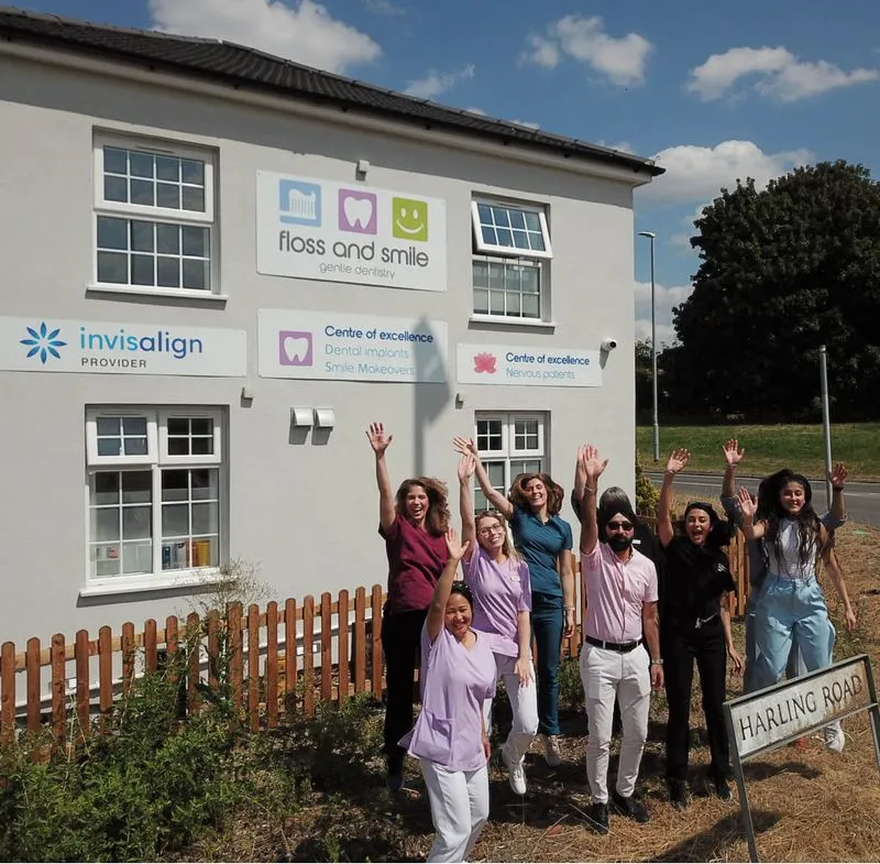 Group of seven cheerful dental clinic staff waving in front of a building with signs for Floss and Smile gentle dentistry and Invisalign provider under a blue sky.