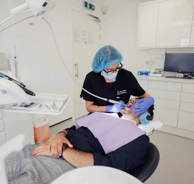 Dentist wearing blue hairnet, mask, and gloves treating a male patient reclining in a dental chair in a modern clinic.