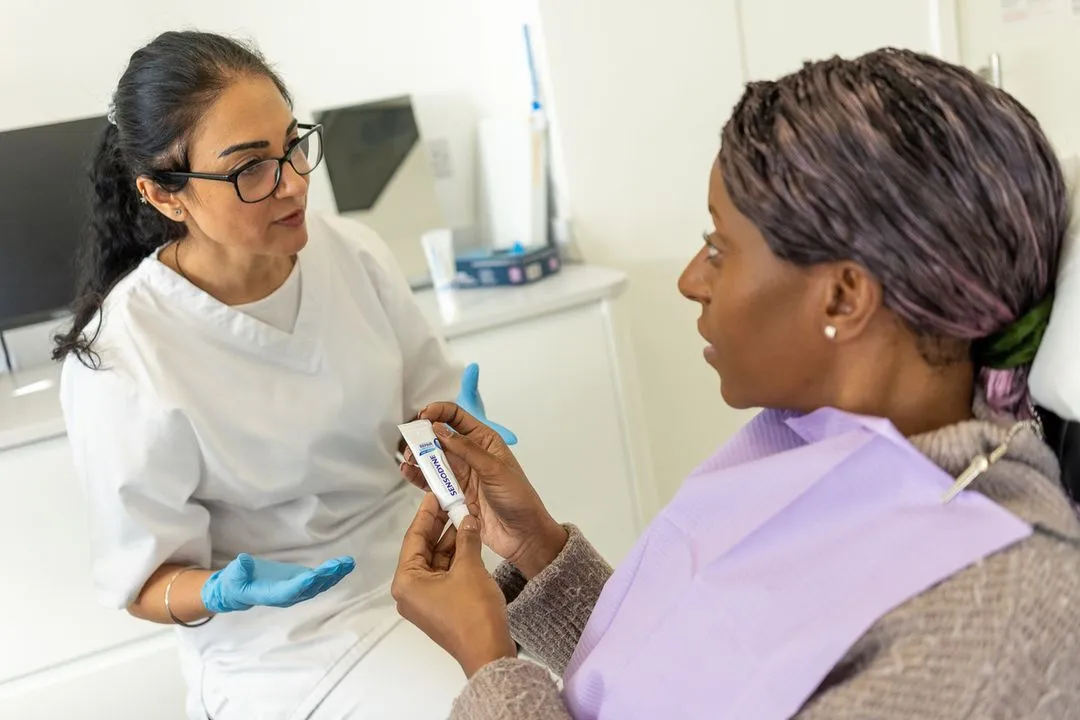 Dental professional in white coat and blue gloves explains product to patient holding Sensodyne toothpaste tube.