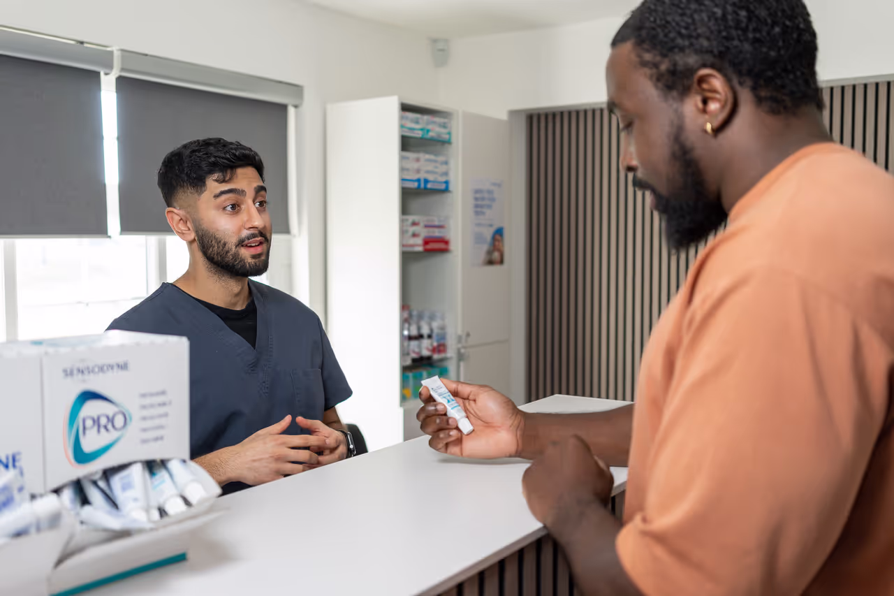 Pharmacist discussing medication with a customer holding a tube of cream at a pharmacy counter.
