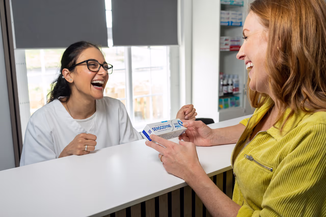 Smiling woman in yellow jacket holding a Sensodyne toothpaste box while conversing with a laughing pharmacist behind the counter.