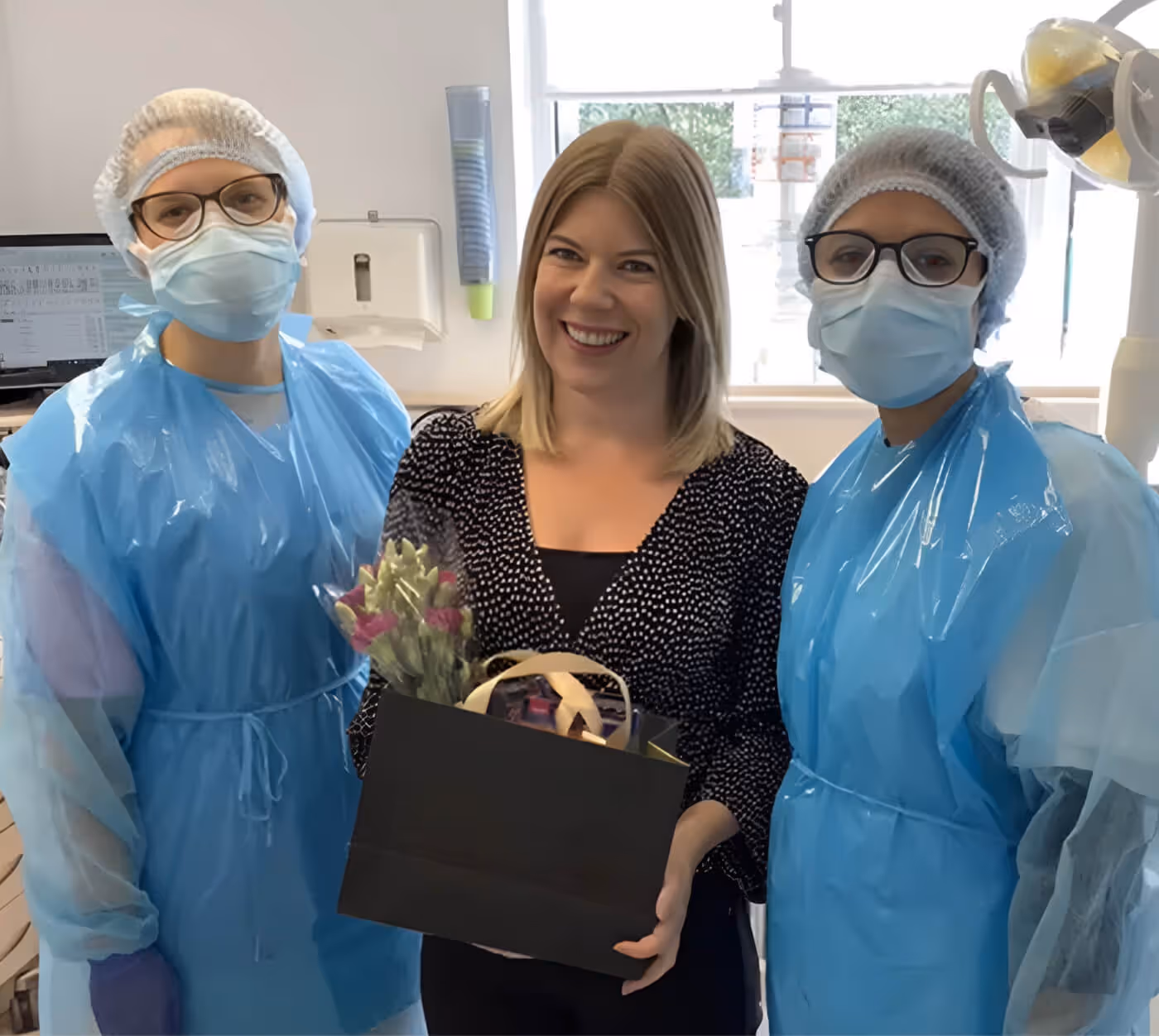 Smiling woman holding a gift bag with flowers stands between two healthcare workers in blue protective gowns, masks, and hair covers in a medical setting.