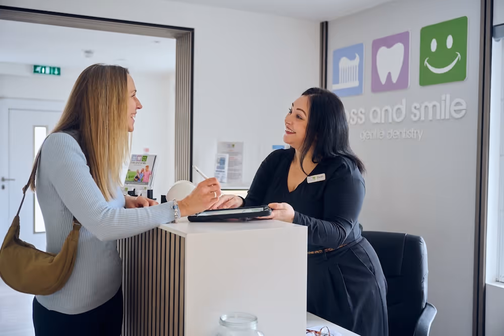 Receptionist smiling and assisting a woman who is signing on a tablet at a dental clinic front desk.