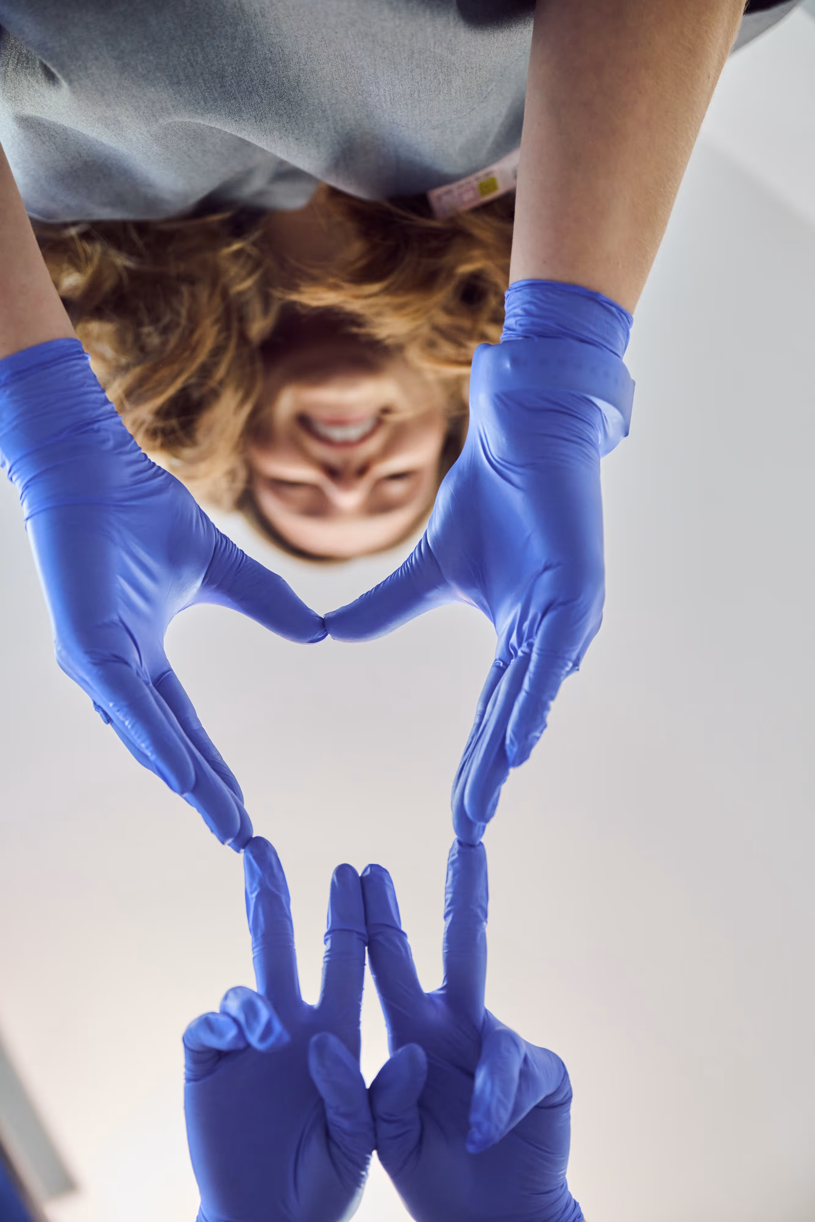 Person wearing blue gloves forming a heart shape with their hands while smiling and looking downwards.