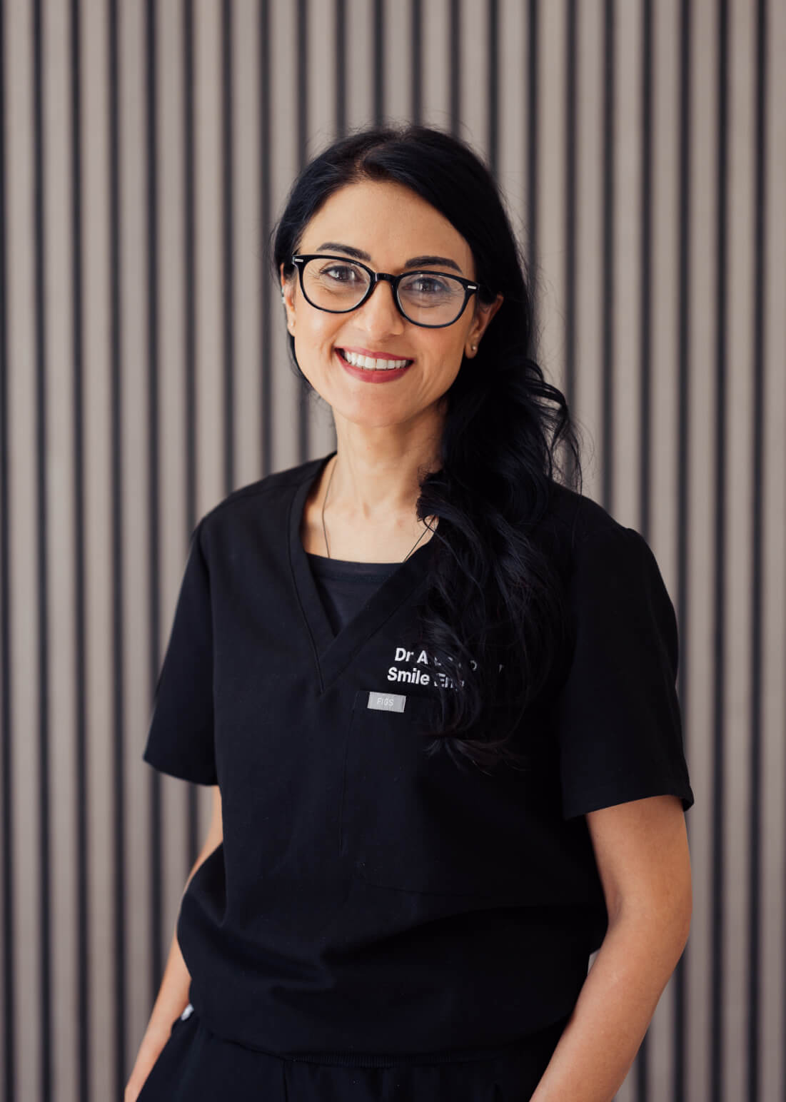 Smiling woman with long dark hair and glasses wearing black medical scrubs in front of a striped background.
