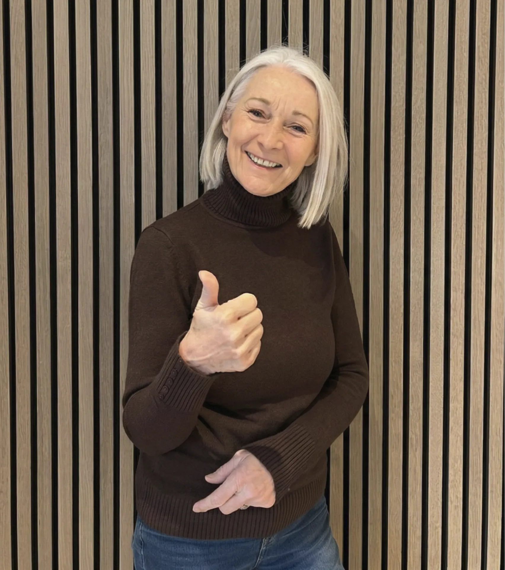 Smiling older woman with gray hair wearing a brown turtleneck sweater and blue jeans giving a thumbs-up gesture.