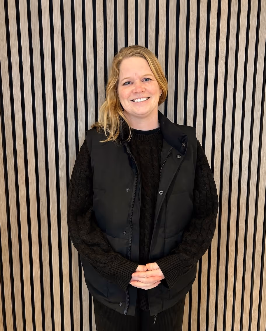 Smiling woman with blonde hair wearing a black sweater and black vest, standing in front of a vertically striped wooden wall.