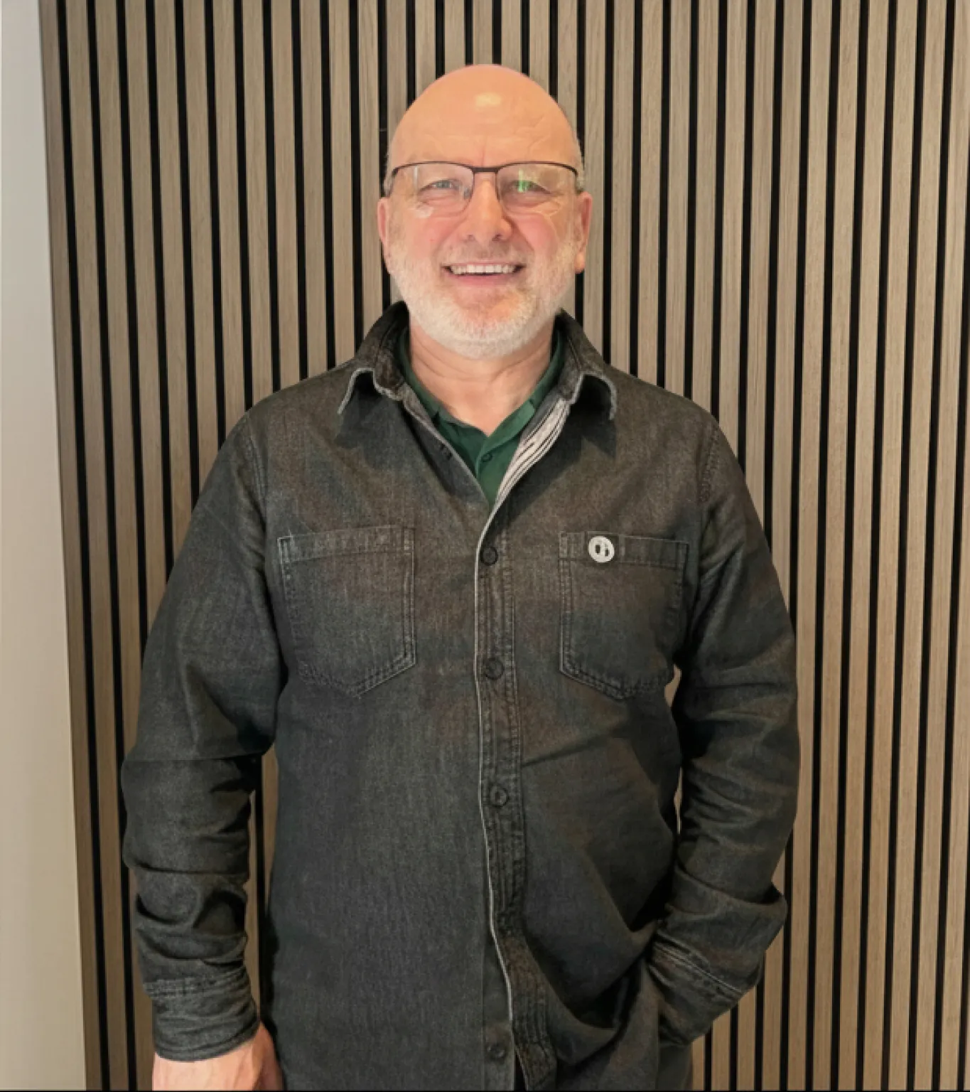 Smiling elderly man with glasses and a beard wearing a dark button-up shirt standing against a vertical wooden slat wall.