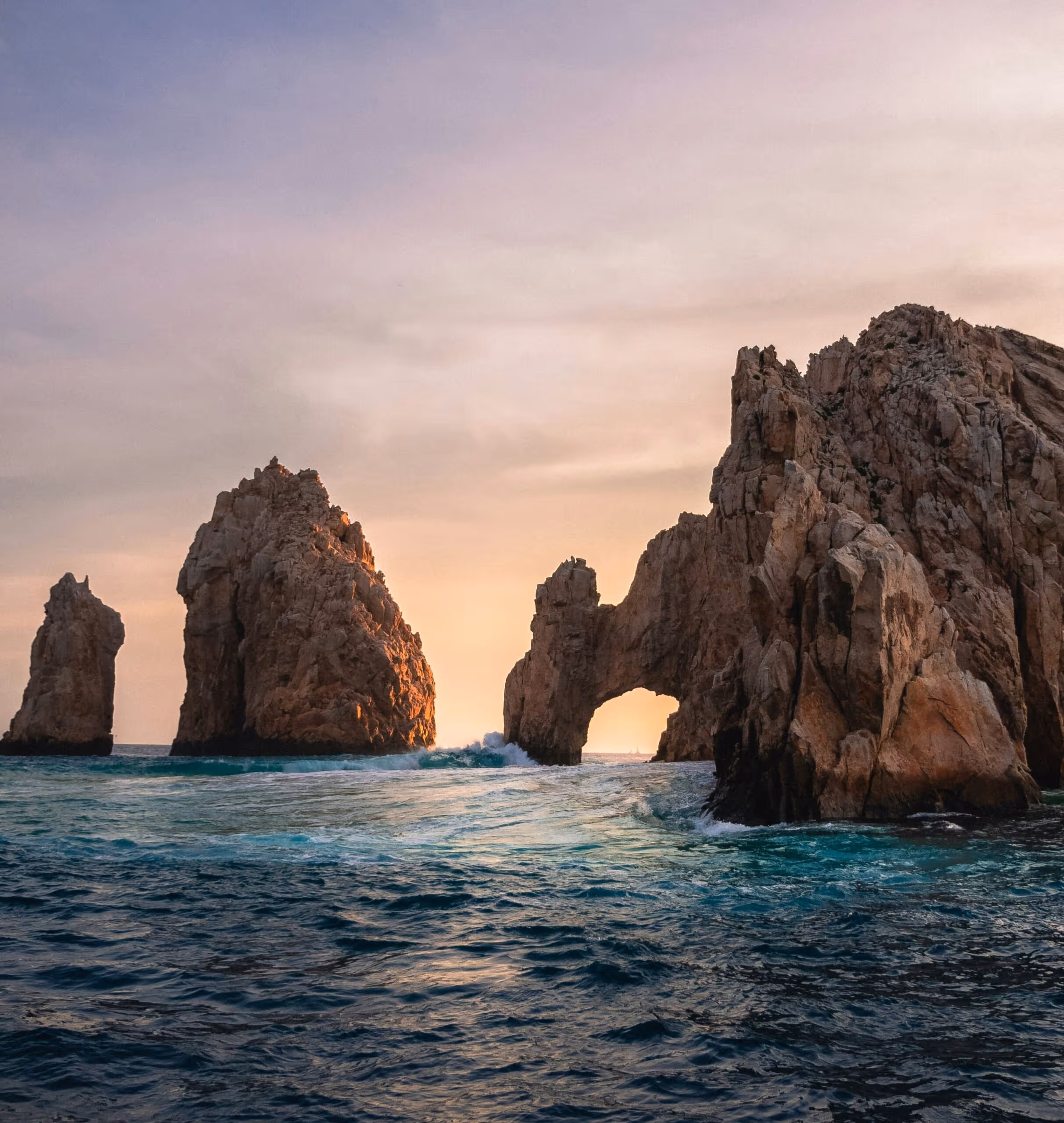 A scenic view of large rock formations emerging from the ocean, with one arch-shaped formation, against the backdrop of a soft sunset sky and gentle ocean waves