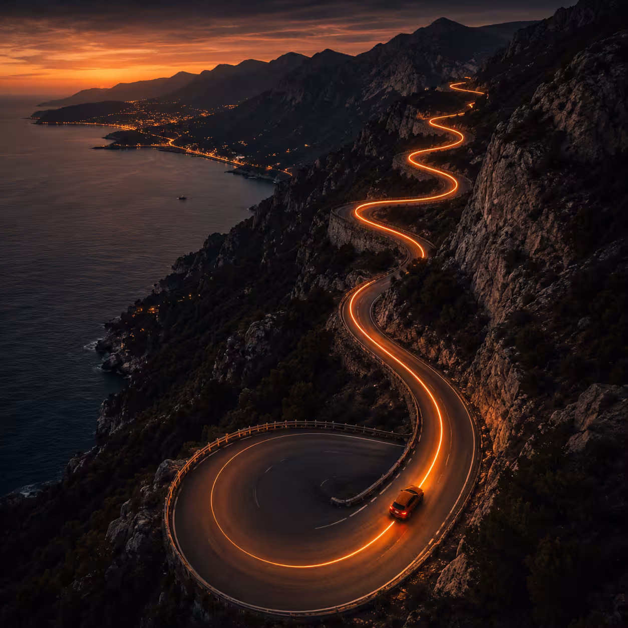 Car driving on winding mountain road at sunset with lights tracing the path along the coast.
