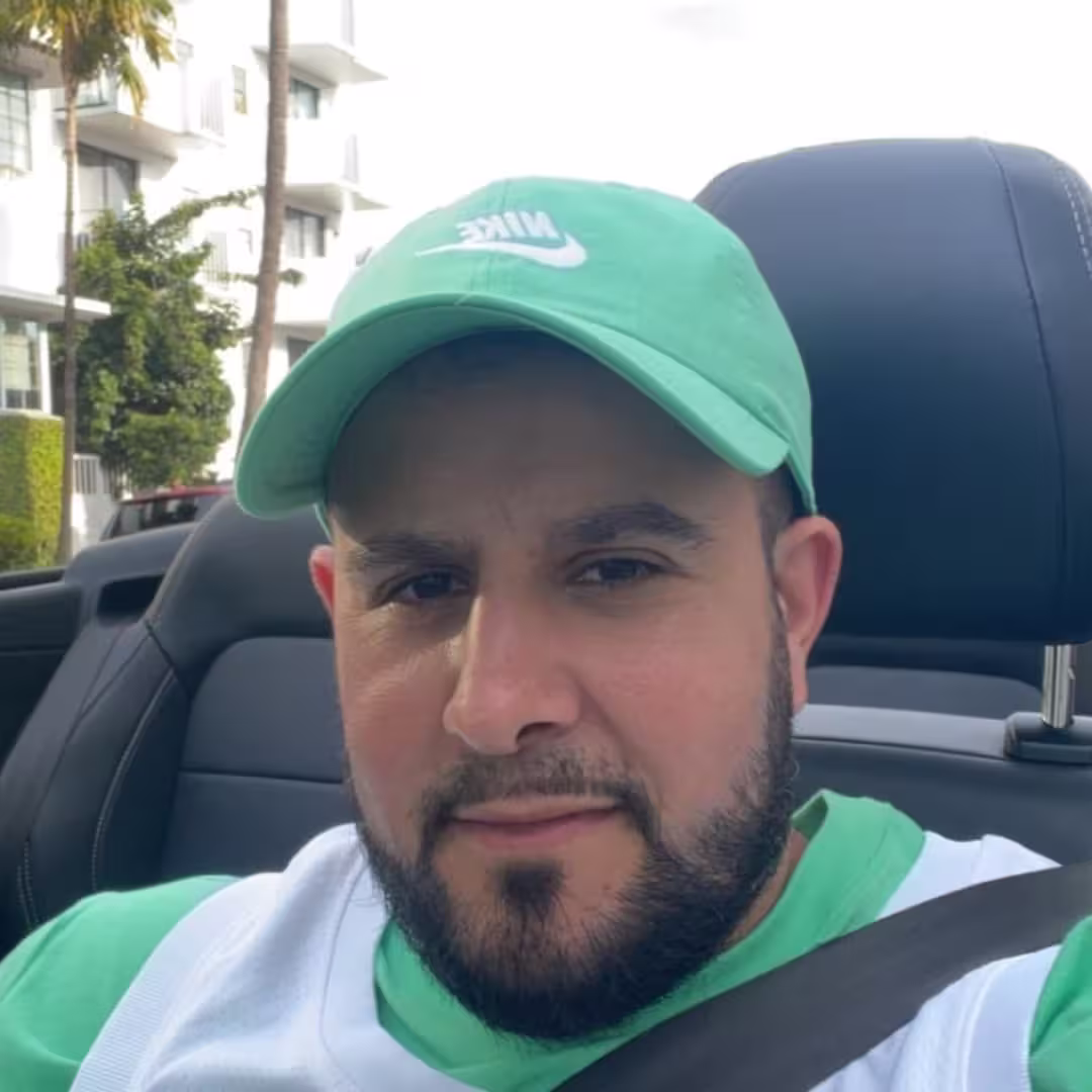 Man with beard wearing green Nike cap and white-green shirt sitting in a car seat with seatbelt on.