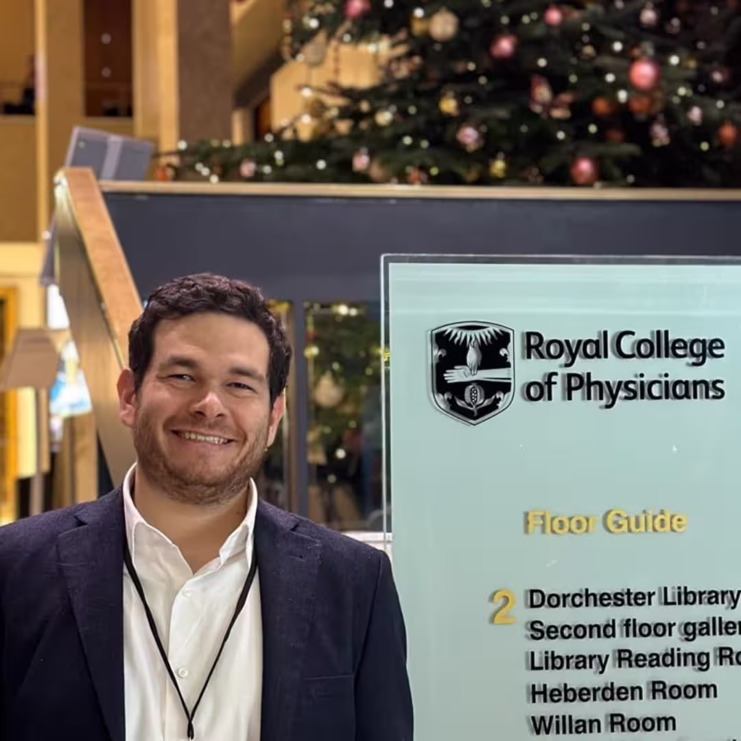 Smiling man in suit standing beside Royal College of Physicians floor guide sign with Christmas decorations behind.