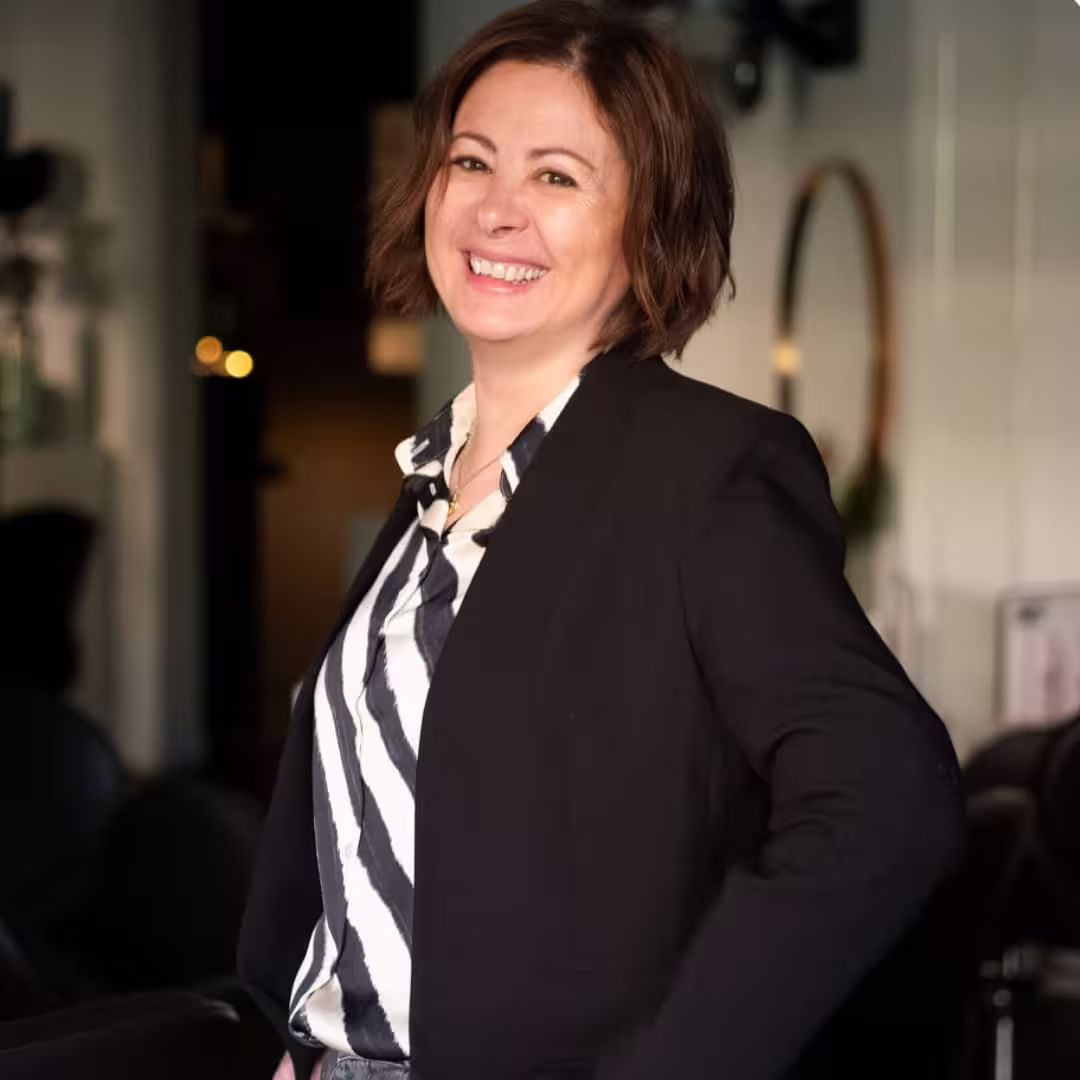 Smiling woman with short brown hair wearing a black blazer and striped shirt indoors.