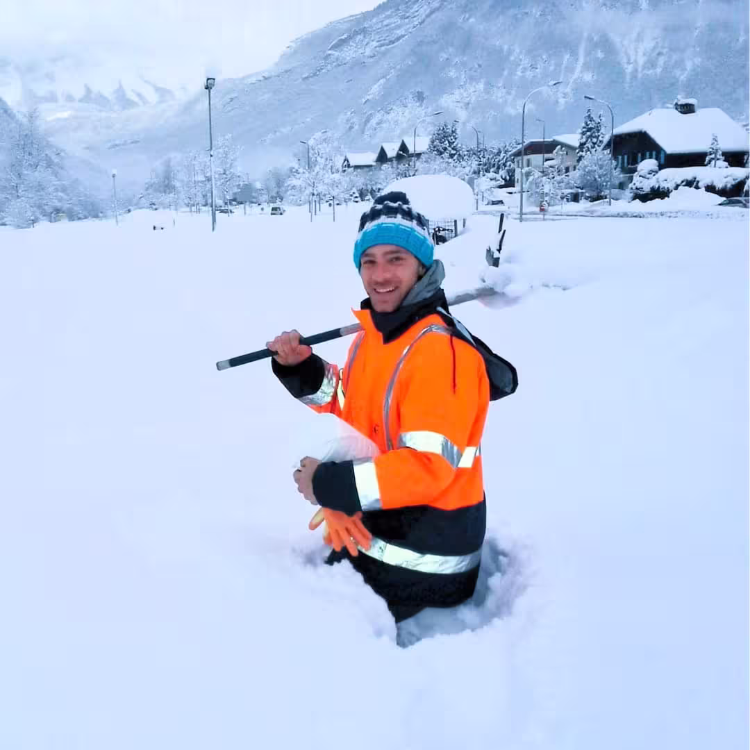 Man in bright orange reflective jacket standing waist-deep in snow holding a shovel, smiling.