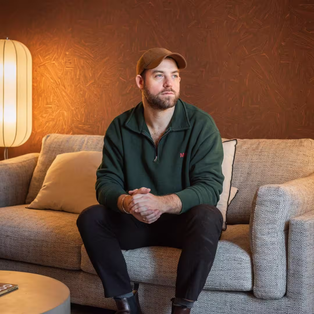 Bearded man in a cap and dark green sweater sitting on a beige couch looking off to the side.