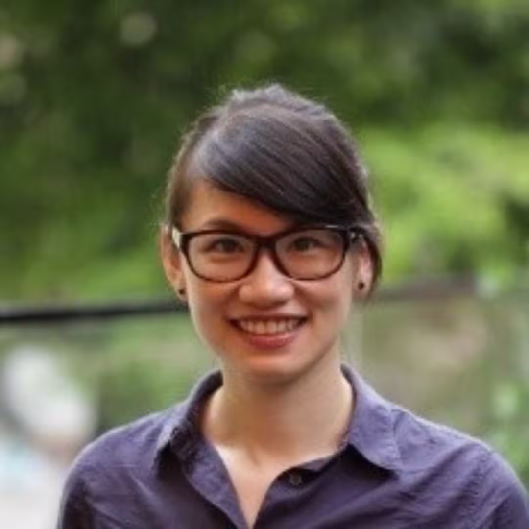 Smiling young woman with glasses and dark hair wearing a purple shirt, standing outdoors with blurred greenery.