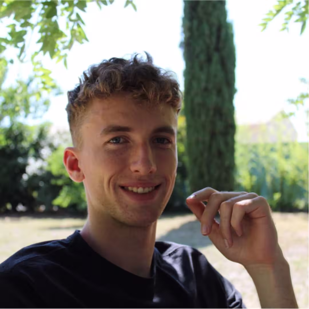 Young man with curly hair smiling outdoors with trees and greenery in the background.