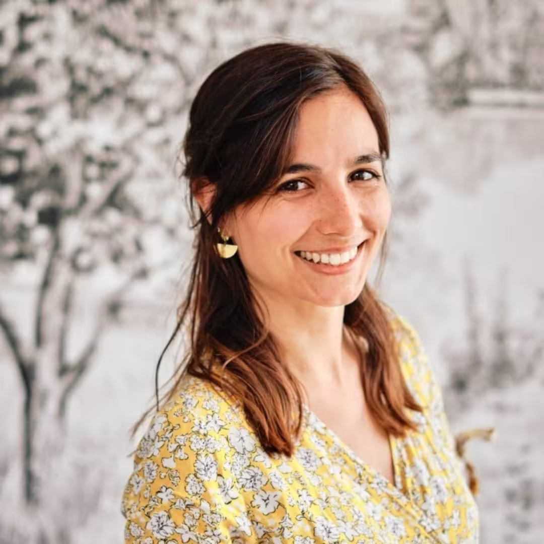Smiling woman with brown hair wearing a yellow floral top and gold earrings.