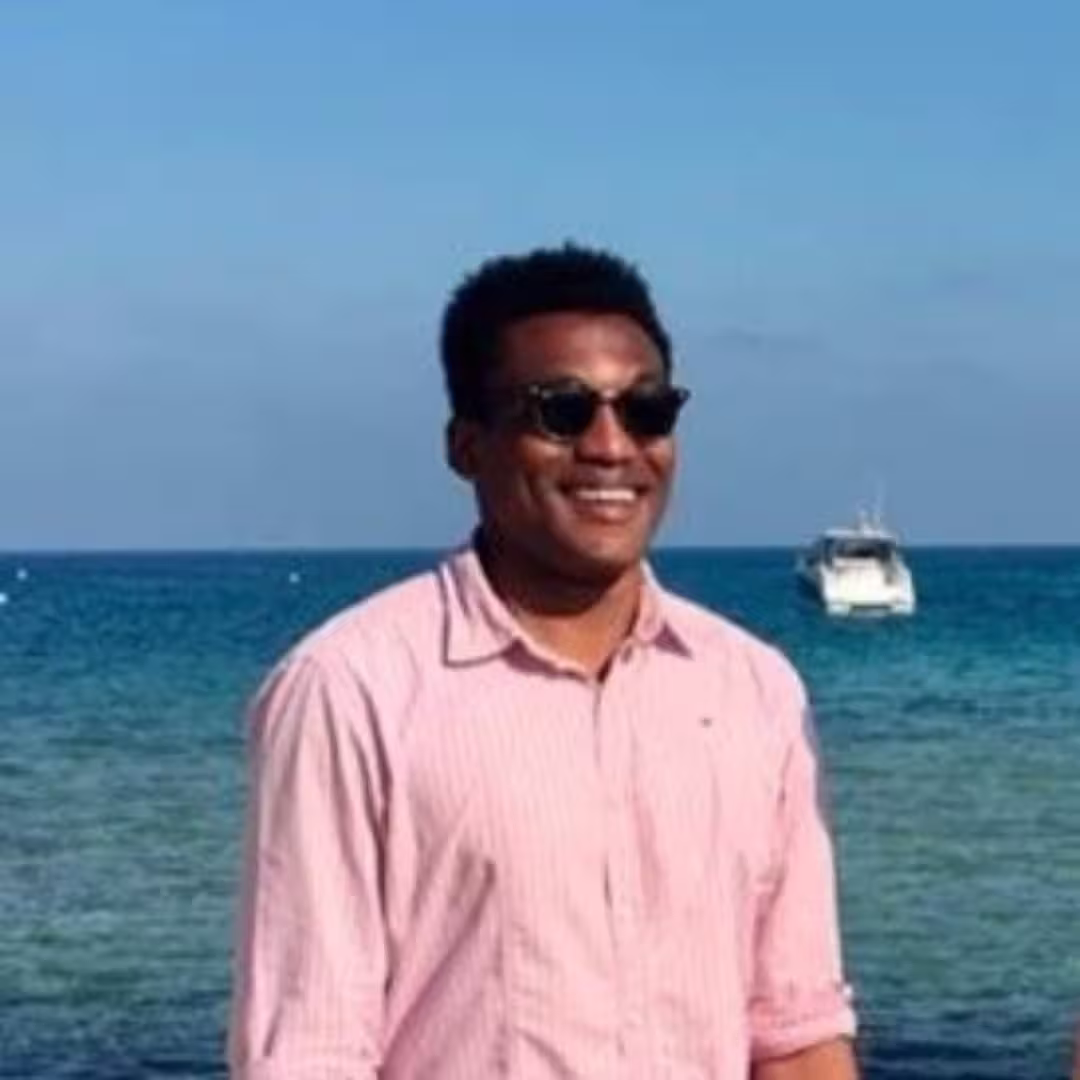 Man in pink shirt and sunglasses smiling with ocean and boat in the background.