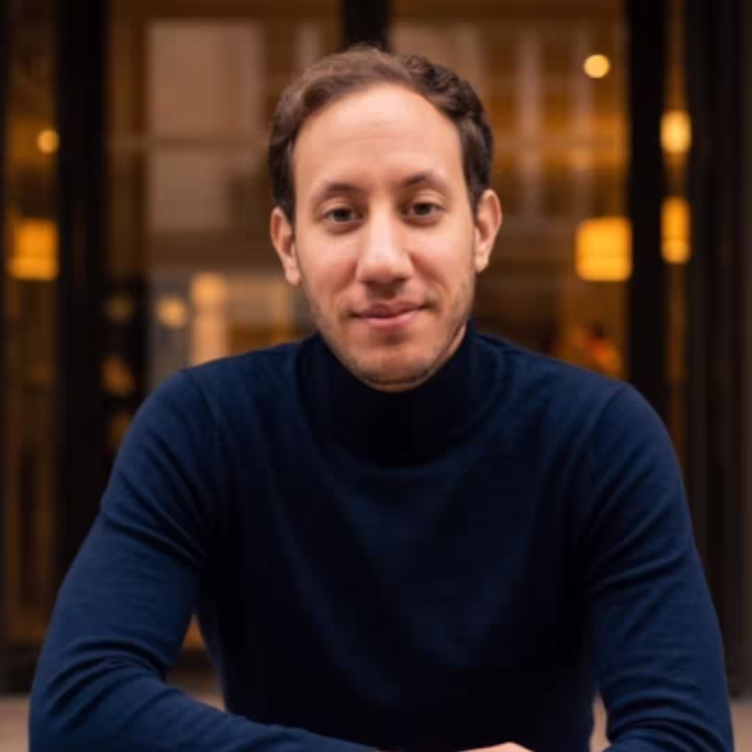 Man with short curly hair wearing a dark blue turtleneck sweater, smiling gently at the camera.