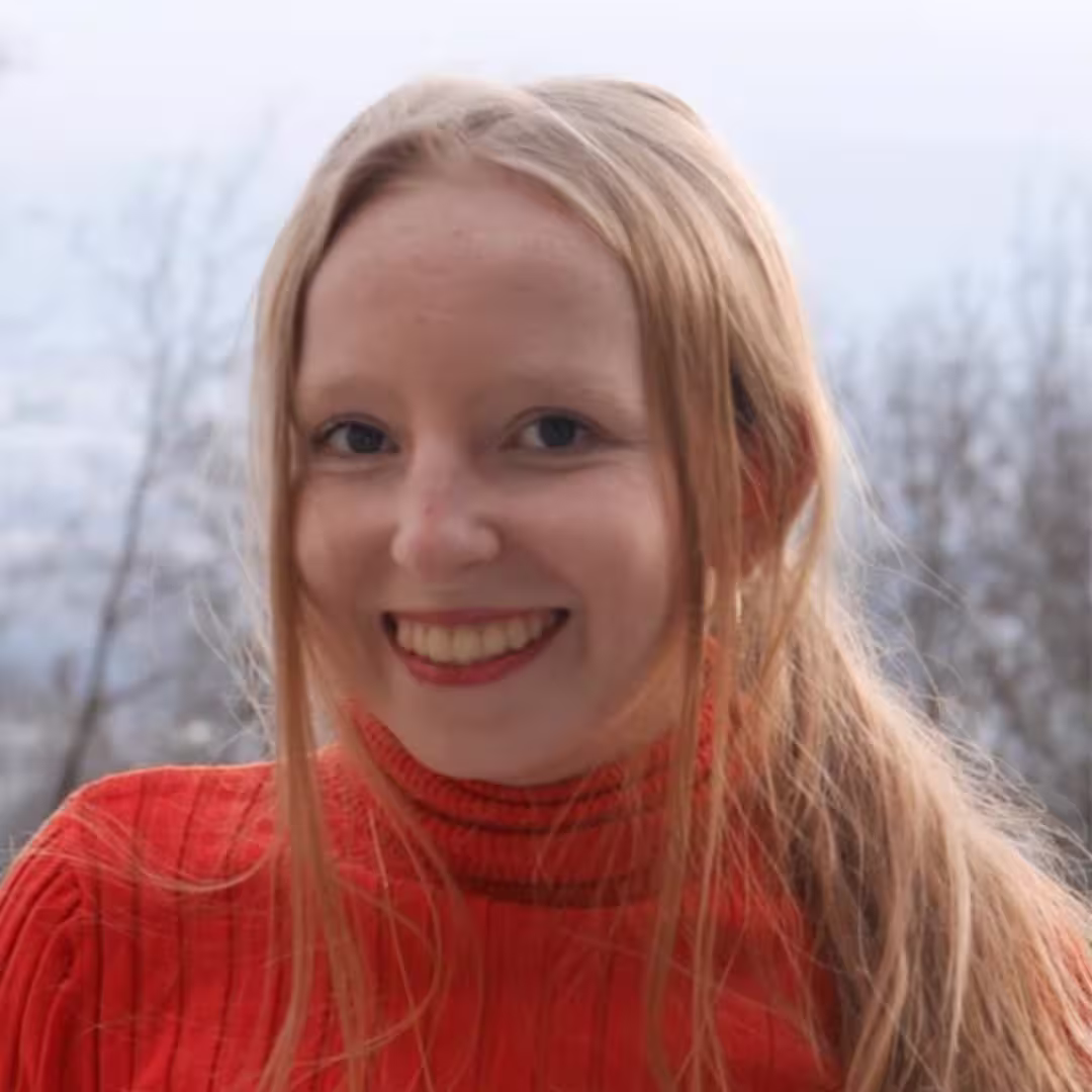 Smiling young woman with long blonde hair in red sweater outdoors with a blurred background.