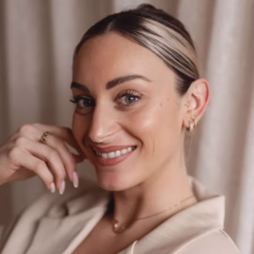 Close-up of a smiling woman with blonde hair pulled back, wearing light makeup and a beige jacket.