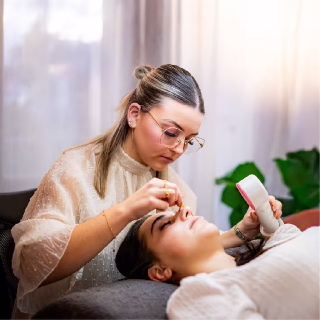 Woman with glasses carefully applying a beauty treatment to a reclining client's face.
