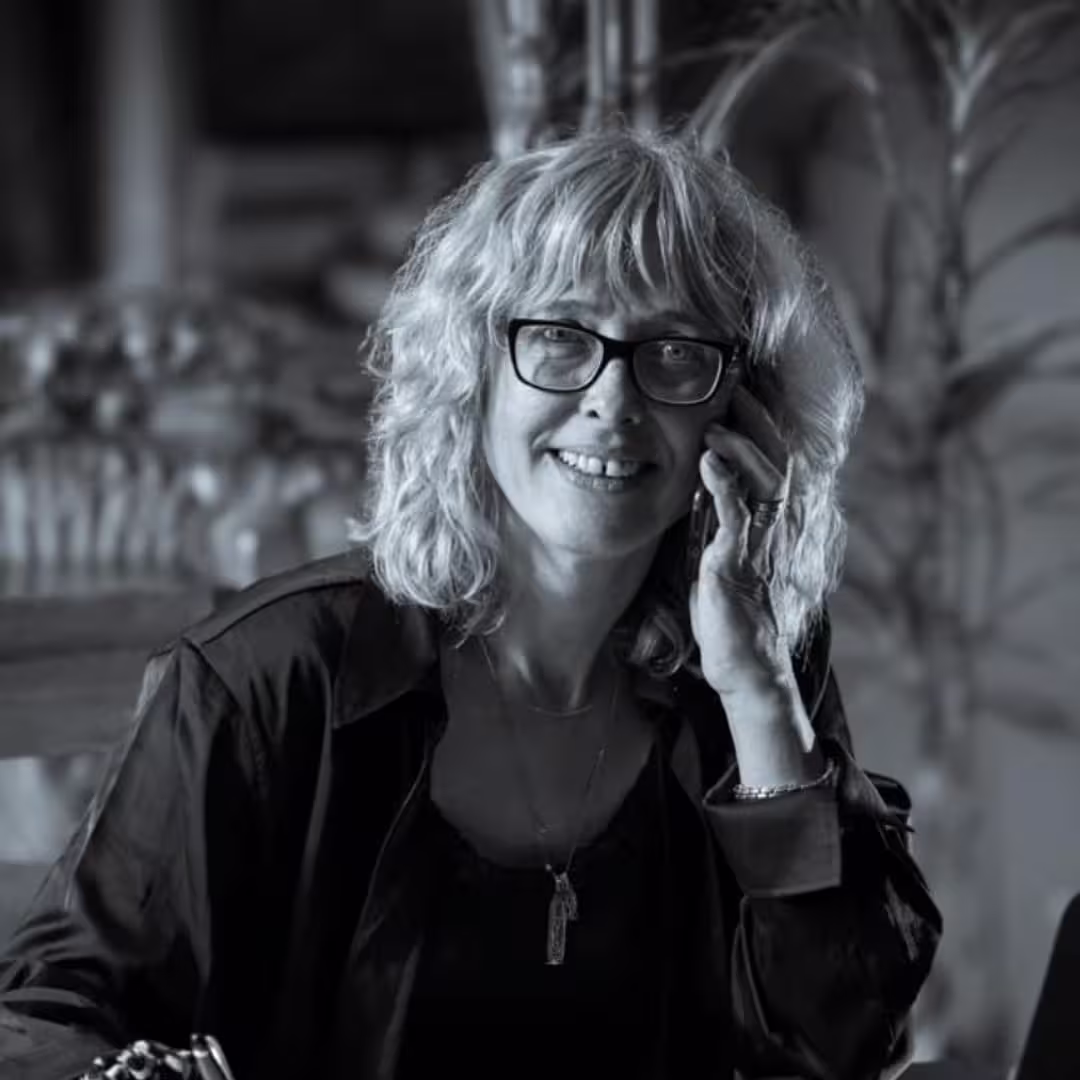Smiling woman with curly hair and glasses talking on a phone in a softly lit room.