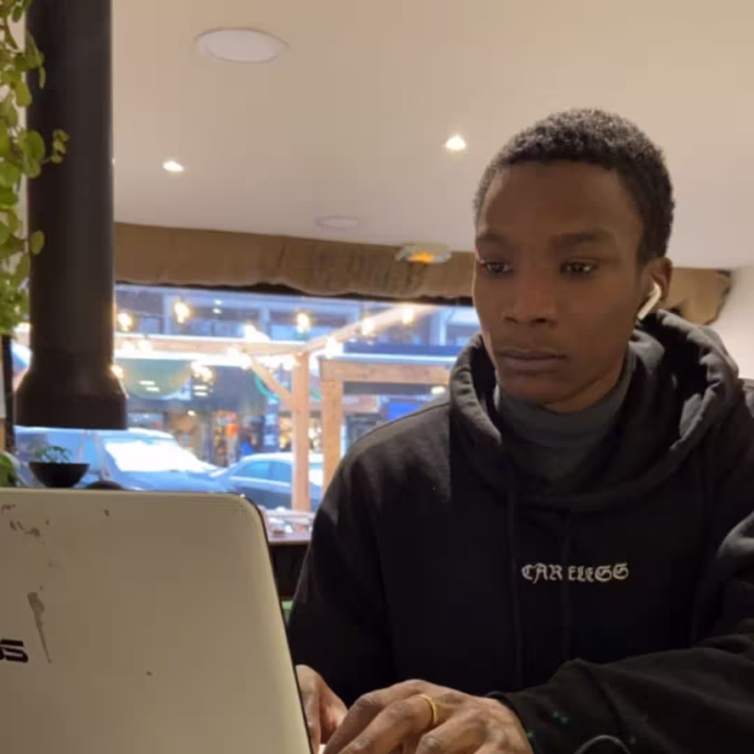 Person wearing earbuds focused on a laptop while seated indoors near a window with street view.