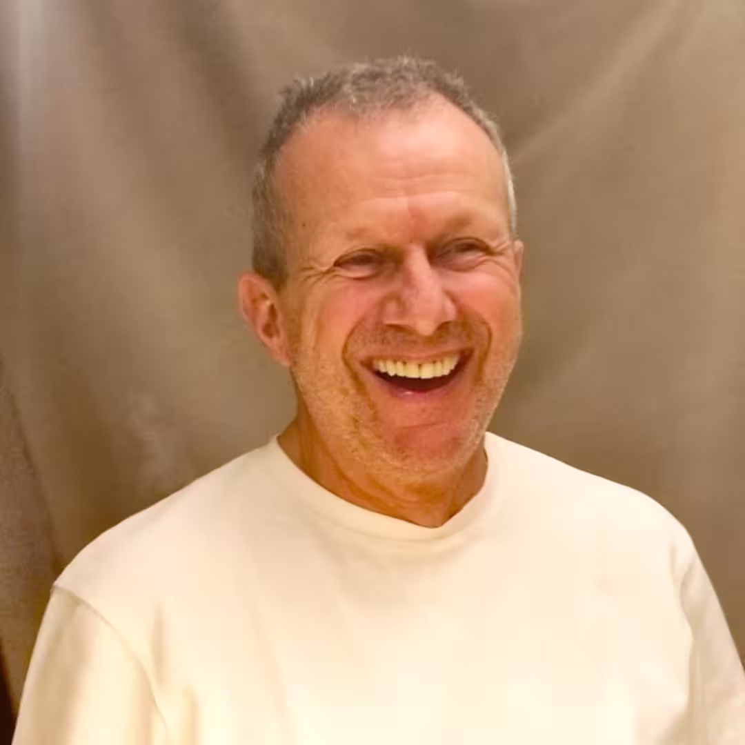 Middle-aged man with short hair and beard smiling wearing a white shirt against a brown background.