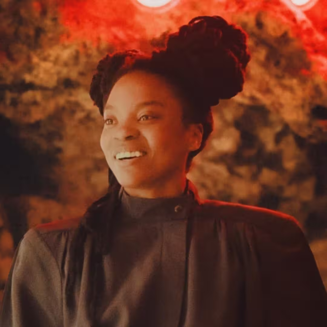 Smiling woman with braided hair styled in large buns wearing a dark high-neck outfit.