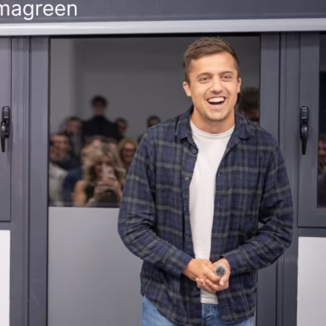 Smiling young man in a plaid shirt standing indoors with a crowd visible behind a glass door.