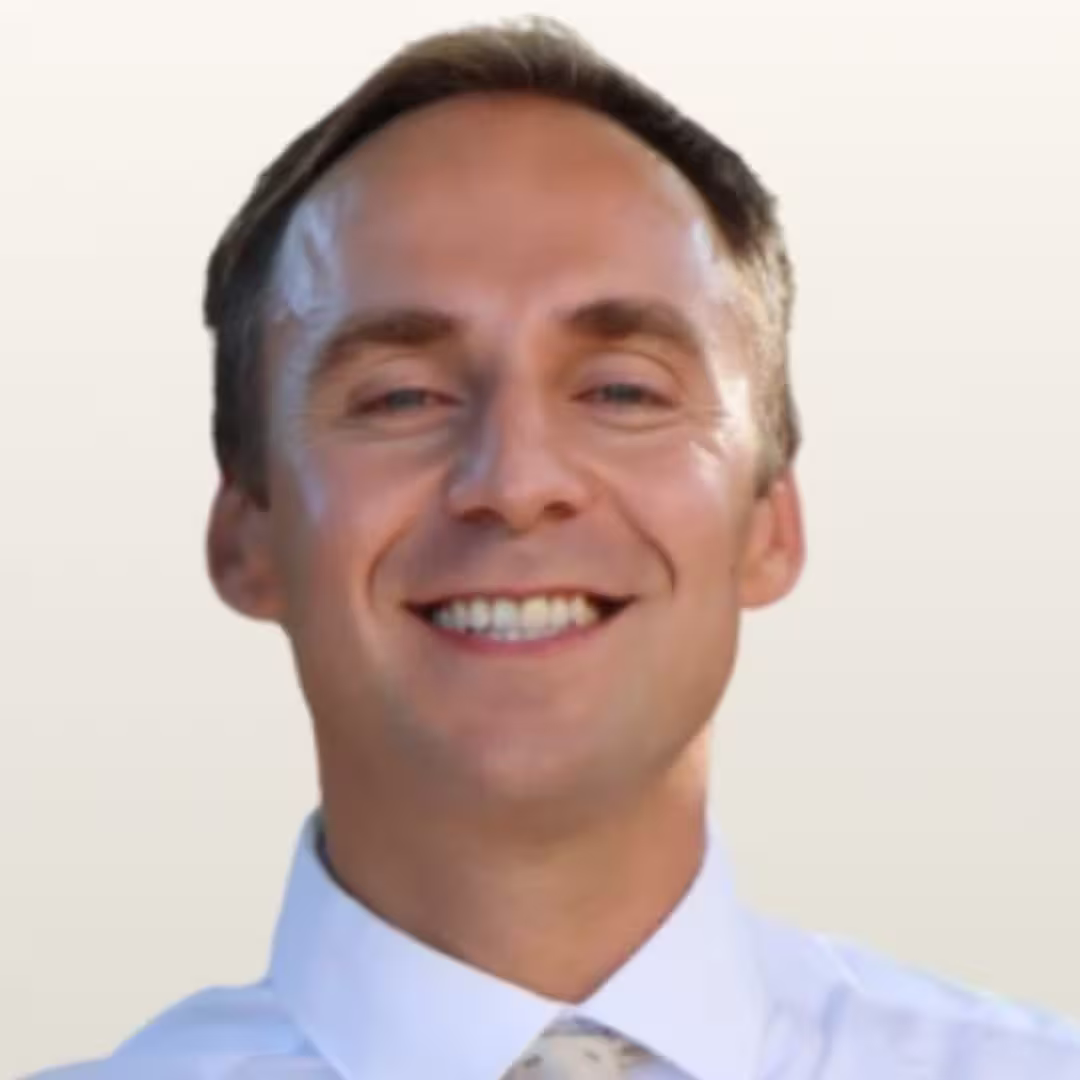 Smiling man with short hair wearing a white shirt and patterned tie on a light background.