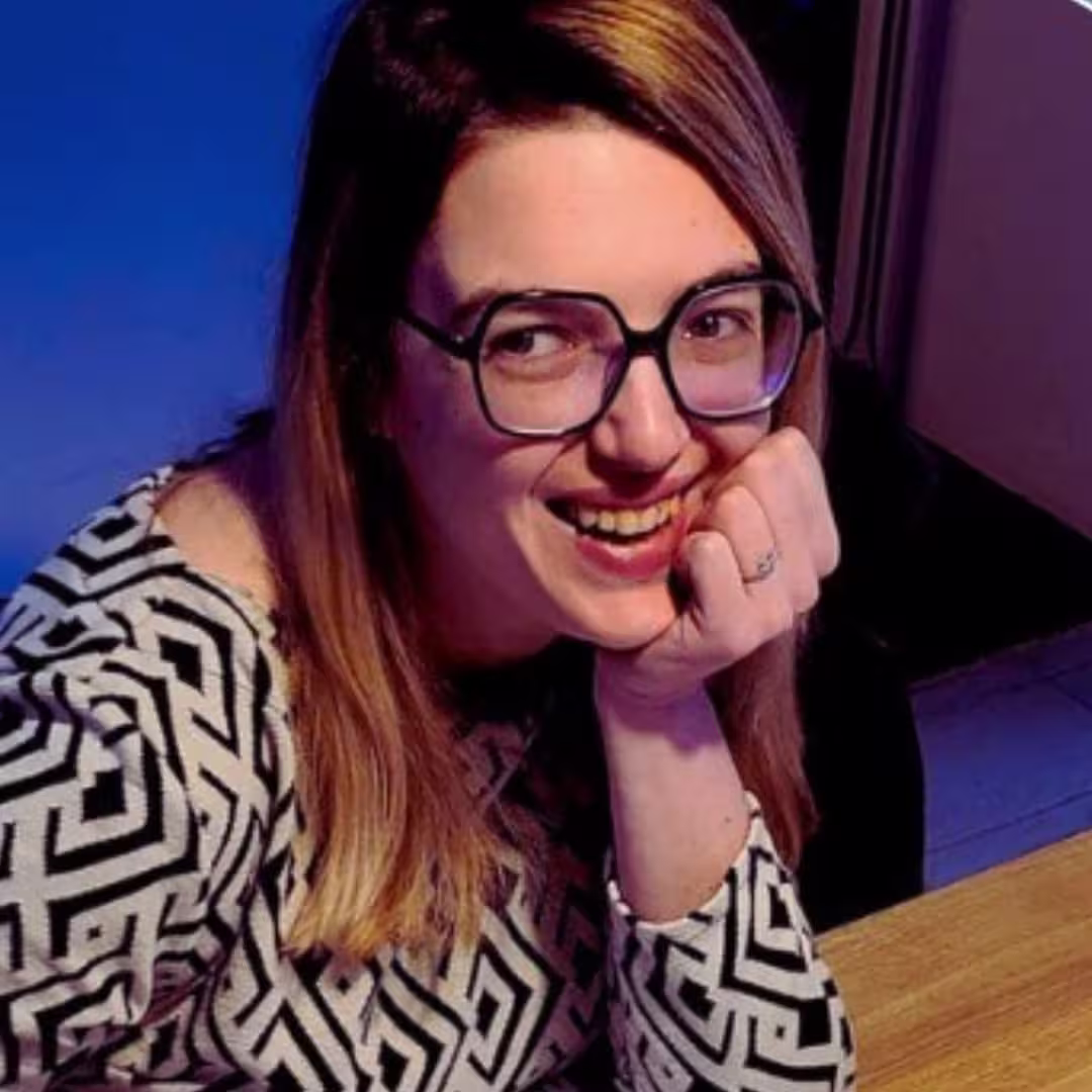 Smiling woman with long hair and glasses in a patterned top resting her chin on her hand.