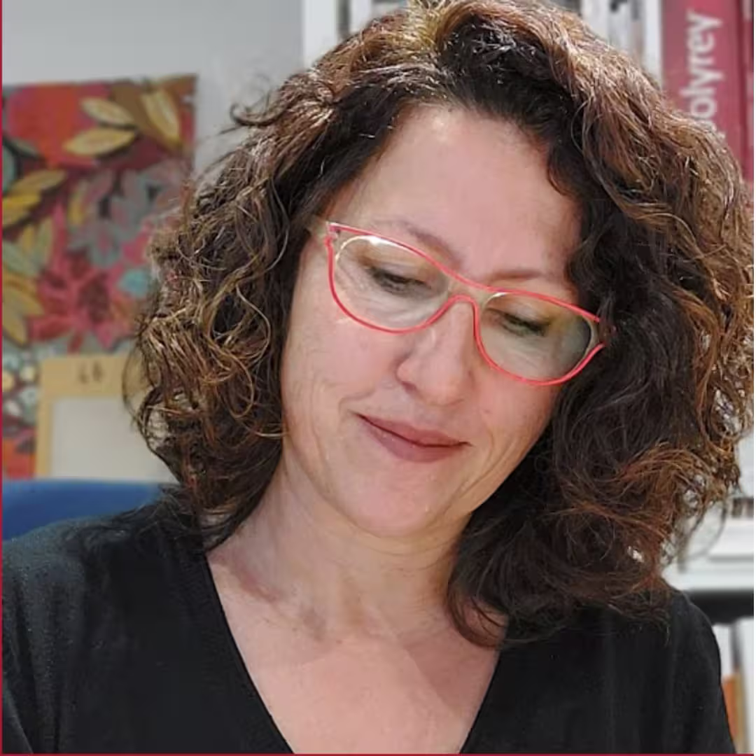 Woman with curly hair and red glasses looking down and smiling softly indoors.