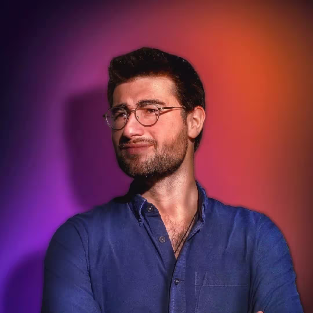 Man with glasses and beard, wearing a blue shirt, standing with arms crossed against a purple and orange background.