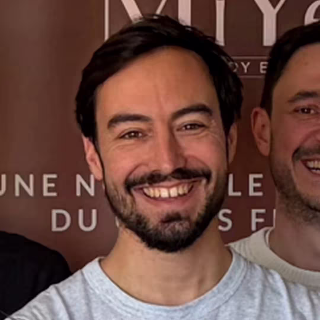 Smiling man with dark hair and beard wearing a light gray shirt, with another person partially visible.