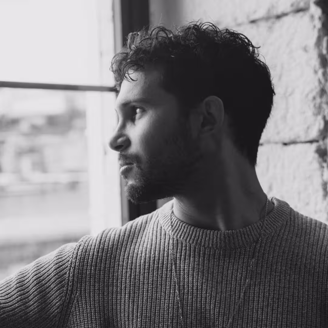 Black and white profile of a man with curly hair and beard looking out a window beside a stone wall.