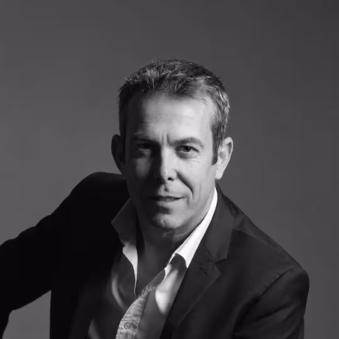 Black and white portrait of a middle-aged man in a blazer and open-collared shirt against plain background.