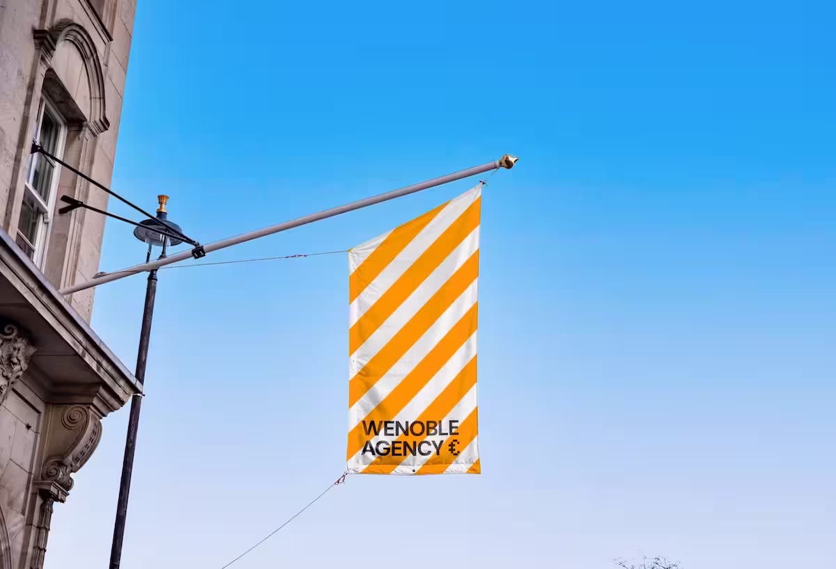 Flag with diagonal orange and white stripes and text ‘Wenoble Agency’ hanging from building.
