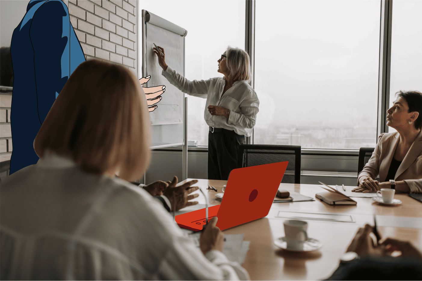 A businesswoman stands pointing at a flip chart while a diverse group of colleagues listen attentively in a modern conference room with large windows. A red laptop is on the table along with coffee cups and notebooks.