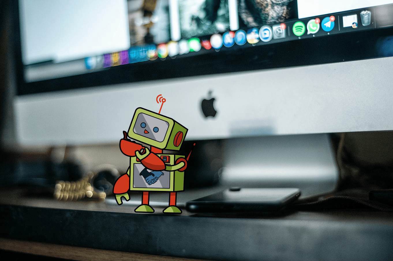 Cartoon green and red robot with smiling face and antenna standing on a desk in front of an Apple iMac computer screen with multiple app icons visible on the screen dock.