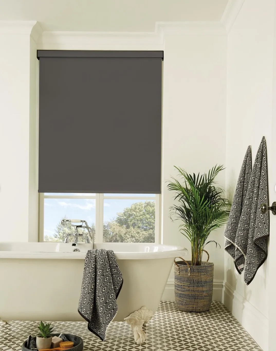 White clawfoot bathtub with black patterned towels, potted plant, and a dark roller blind covering the window.